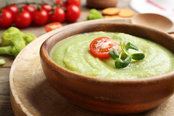 Bowl of broccoli cream soup with tomato and microgreens served on wooden tray, closeup